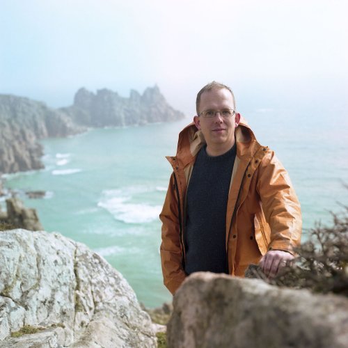 Writer, Wyl Menmuir, standing on cliff with sea view in the background.