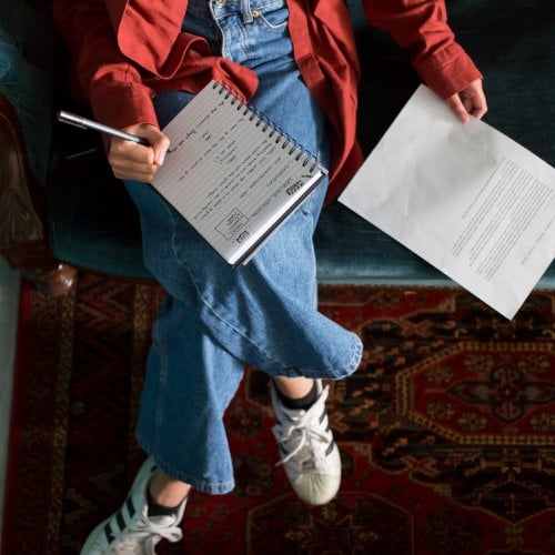 An aerial photo of a girl sat on a sofa with a notebook on her lap and a piece of paper next to her 