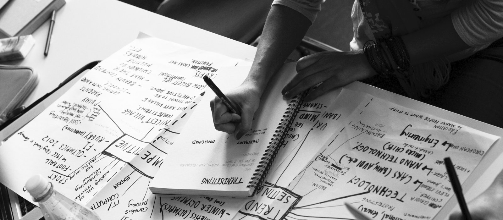 Grayscale image of students hands writing and filling pages in front of them with ideas