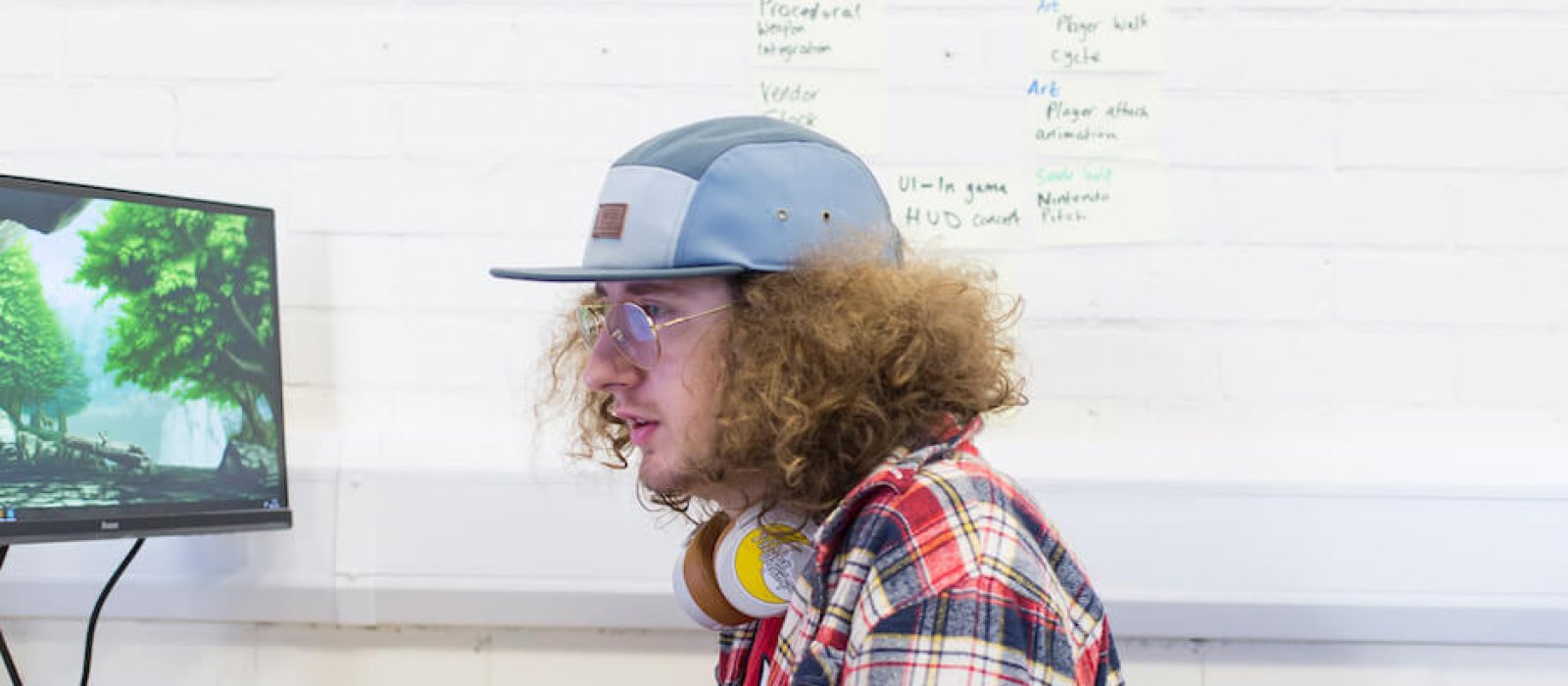 Curly haired male games student with retro glasses and cap sat at computer