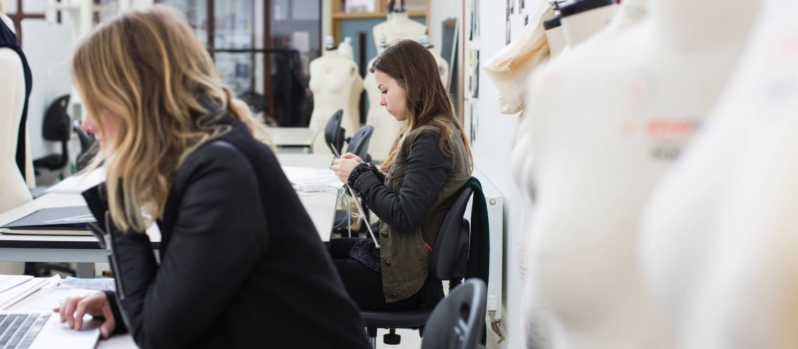 Two students in fashion studio, one on laptop and one knitting.