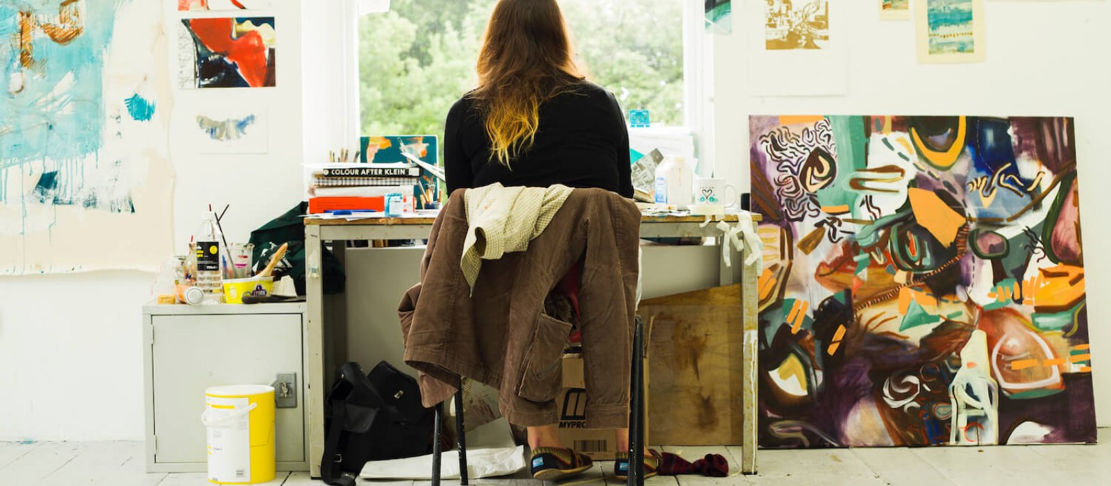 Back of student sitting at desk in front of window surrounded by artwork and paintings.