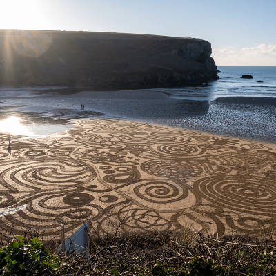 Photo of Mawgan Porth beach with the sun setting over the headland and a series of swirling patterns drawn into the sand