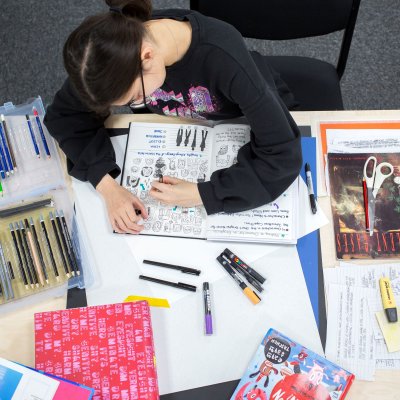 A female student drawing in a sketchbook surrounded by stationary