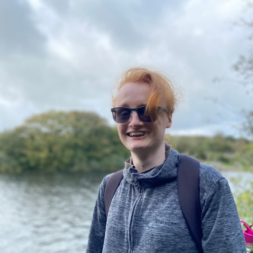 A Falmouth University students next to a water and trees, wearing a hoodie and sunglasses