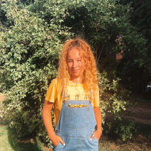 A Falmouth University student wearing denim dungarees and standing near bushes