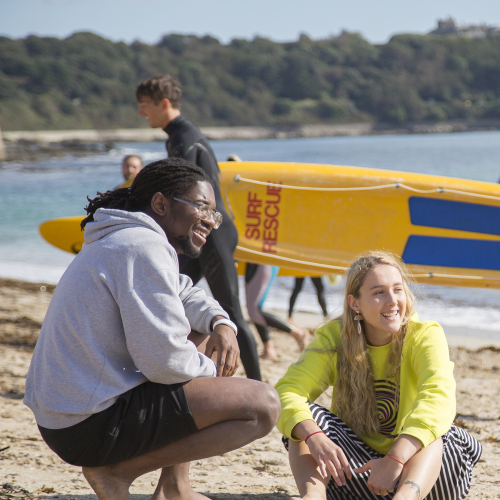 Two students sitting on the beach in front of a yellow surf rescue board
