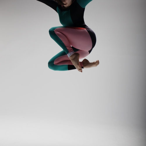 Dancer jumping high with arms spread and legs tucked under in grey backdrop studio.