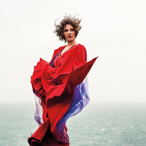Model posing by sea, red outfit and hair blowing in the wind.