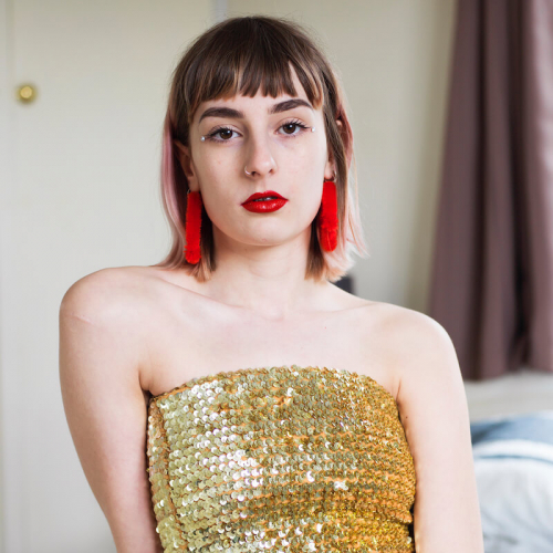 Model with gold sequin top and red tassel earring and red belt sat on a bed.