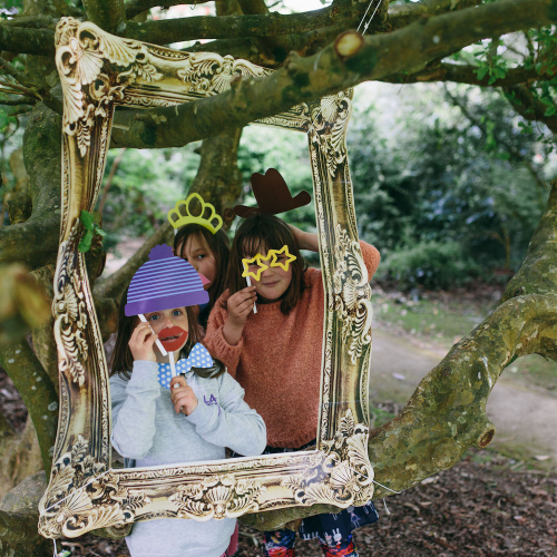 Children posing in woods behind gold gilt picture frame with masks at Little Fay Festival