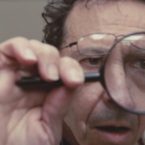 Film still, close up of a male face looking through a magnifying glass.