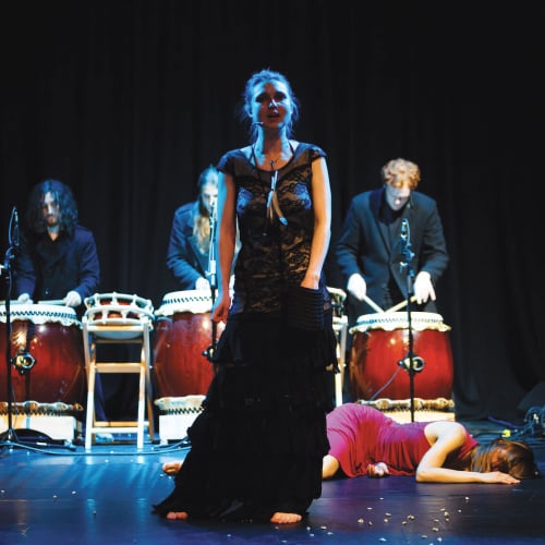 Actor in long black dress, body on the stage floor behind her and 3 drummers at the back of the stage.