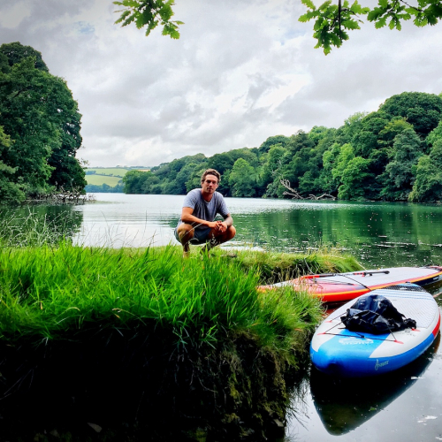 Falmouth University student crouching on the banks of the river with trees and paddleboards