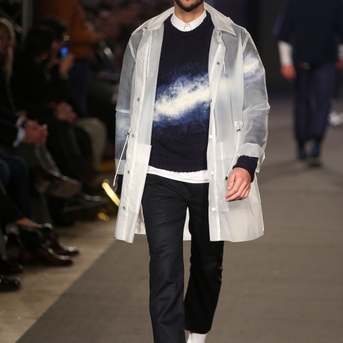Male walking a catwalk, navy outfit, white overcoat and white flash across the front.