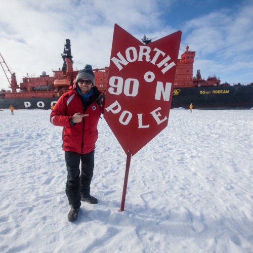 A man next to the North Pole sign with ship behind
