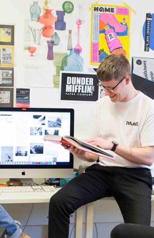 Three Falmouth University students sat round an iMac with graphic design posters on the wall