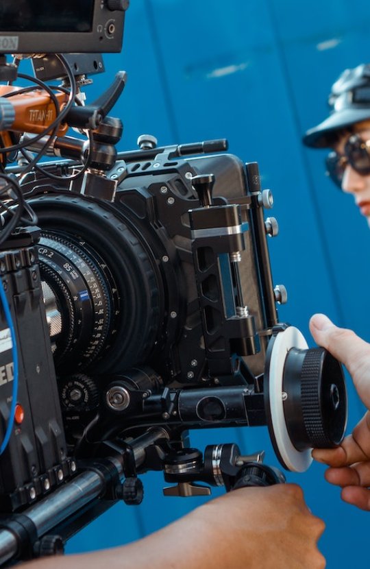 A man controlling a RED camera filming a women wearing sunglasses, a leather hat and jacket